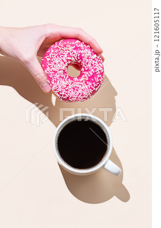 Hand holding fresh pink donut on beige background with shadow and cup of espresso coffee. 121605117