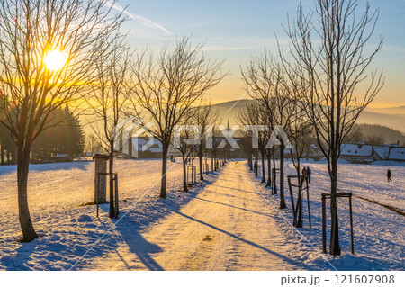 Golden light of sunset bathes the snow-covered path in Prichovice, Czechia. Bare trees line the way, leading toward distant hills and quaint buildings in the serene winter scene. Golden light of sunset bathes the snow-covered path in Prichovice, Czechia. Bare trees line the way, leading toward distant hills and quaint buildings in the serene winter scene. 121607908