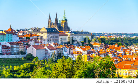 The grand architecture of Prague Castle towers above the vibrant rooftops of the city. Lush greenery complements the lively urban landscape, creating a picturesque view on a clear day. The grand architecture of Prague Castle towers above the vibrant rooftops of the city. Lush greenery complements the lively urban landscape, creating a picturesque view on a clear day. 121608431