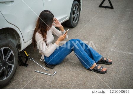 A woman dressed in jeans is struggling with car trouble while using her phone urgently A woman dressed in jeans is struggling with car trouble while using her phone urgently 121608695