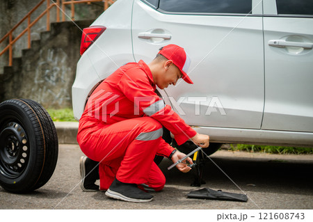 A mechanic skillfully changing a tire on a white car while wearing a bright red uniform 121608743