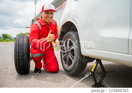 A mechanic is changing a flat tire with a jack on a sunny day, showing expertise 121608763