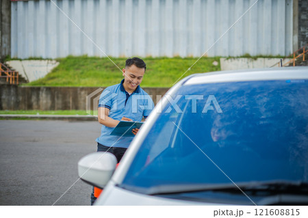 A man is writing on a clipboard by his parked car, completing his checklist 121608815
