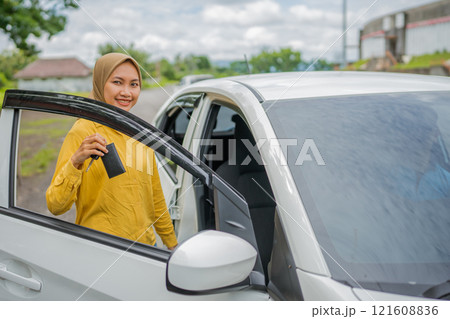A smiling woman confidently holds a car key next to her white vehicle in daylight 121608836