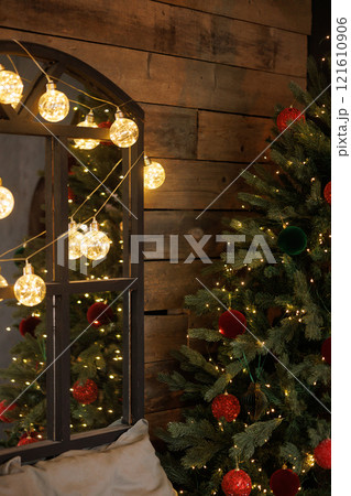 A dark-framed mirror is decorated with a garland of large balls next to a Christmas tree 121610906