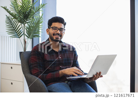 Smiling young Indian man sitting in office chair holding laptop. Casual work environment with natural light. Comfortable and relaxed atmosphere. Modern workspace with greenery. Smiling young Indian man sitting in office chair holding laptop. Casual work environment with natural light. Comfortable and relaxed atmosphere. Modern workspace with greenery. 121617671