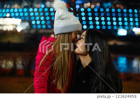 Two people share a warm moment outdoors with colorful lights in the background during a chilly evening 121624958