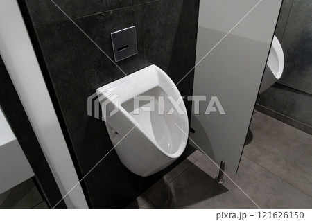 A modern urinal is mounted on a dark tiled wall in a well-lit public restroom. The minimalistic design emphasizes clean lines, creating a contemporary feel in the space A modern urinal is mounted on a dark tiled wall in a well-lit public restroom. The minimalistic design emphasizes clean lines, creating a contemporary feel in the space 121626150