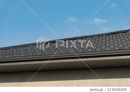 A close-up view of a contemporary residential rooftop is captured under a vibrant blue sky. The sleek metallic tiles reflect sunlight, accentuating the modern architectural design of the home A close-up view of a contemporary residential rooftop is captured under a vibrant blue sky. The sleek metallic tiles reflect sunlight, accentuating the modern architectural design of the home 121626204