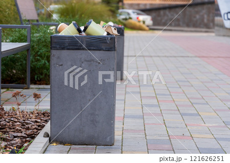 A concrete garbage bin filled to capacity with trash bags sits on a bustling city sidewalk, surrounded by plants and pavers under a clear sky. People pass by, highlighting urban waste challenges 121626251