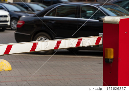 A closed automatic barrier secures the entrance to a parking lot, preventing unauthorized access. The scene captures the red and white striped gate in operation on a busy road A closed automatic barrier secures the entrance to a parking lot, preventing unauthorized access. The scene captures the red and white striped gate in operation on a busy road 121626287