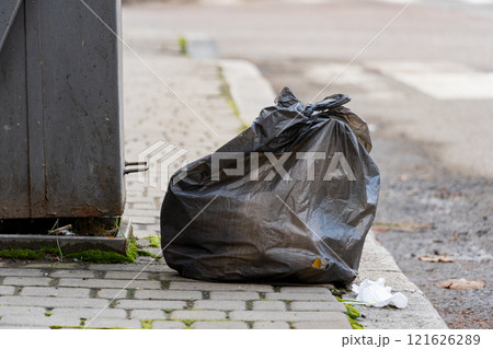 A black garbage bag is placed beside a recycling container on a city street. Litter can be seen on the pavement, highlighting issues of waste management in urban areas 121626289