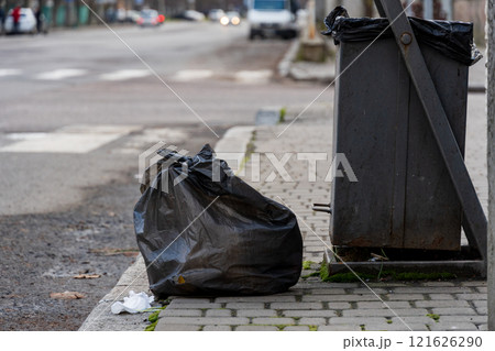 Two black trash bags rest beside a metallic garbage container along a city sidewalk, emphasizing the ongoing issues with urban waste disposal and environmental conservation efforts Two black trash bags rest beside a metallic garbage container along a city sidewalk, emphasizing the ongoing issues with urban waste disposal and environmental conservation efforts 121626290