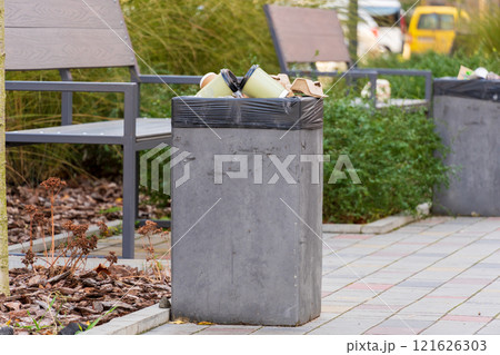 A concrete garbage bin brimming with trash bags and recyclable materials is placed in an outdoor setting. Surrounding benches and greenery create a casual atmosphere A concrete garbage bin brimming with trash bags and recyclable materials is placed in an outdoor setting. Surrounding benches and greenery create a casual atmosphere 121626303