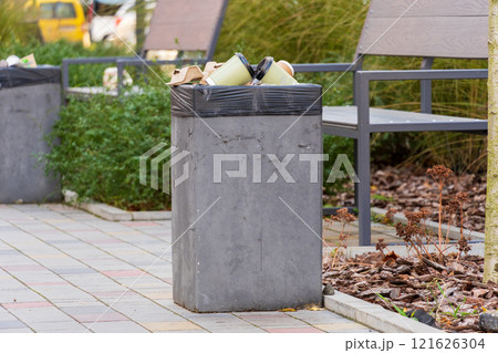 A concrete garbage container is overflowing with trash bags and recyclables in a well-maintained urban park. Nearby benches and greenery enhance the outdoor atmosphere, highlighting waste management 121626304