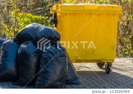 A yellow garbage container sits beside several black trash bags on a paved surface outdoors. The scene captures the importance of proper waste disposal and recycling efforts in the community 121626364