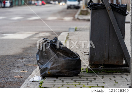 A black garbage bag sits on the curb beside a recycling bin, illustrating daily urban waste management efforts. The setting captures a typical city street with vehicles passing by 121626369