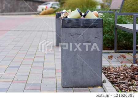 A concrete garbage container is filled with trash bags on a busy city sidewalk. Nearby plants and benches are visible, highlighting urban waste management challenges 121626385