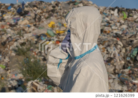 A worker in protective clothing stands amid a large landfill filled with mountains of garbage, visibly engaged in assessing the pollution and its effects on the ecosystem 121626399