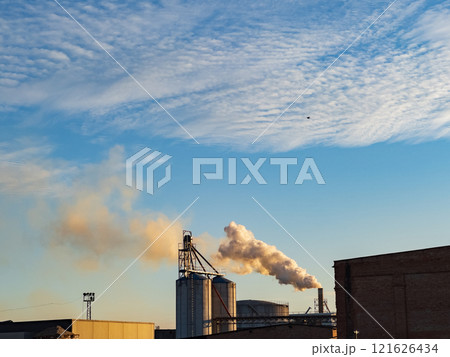 Billowing smoke emerges from smokestacks at an industrial facility during sunset, casting a cloudy backdrop over the manufacturing buildings and machinery Billowing smoke emerges from smokestacks at an industrial facility during sunset, casting a cloudy backdrop over the manufacturing buildings and machinery 121626434