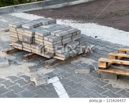 Sturdy concrete pavers are stacked on wooden pallets, surrounded by loose sand and gravel at an urban construction site near a paved street, ready for installation 121626437