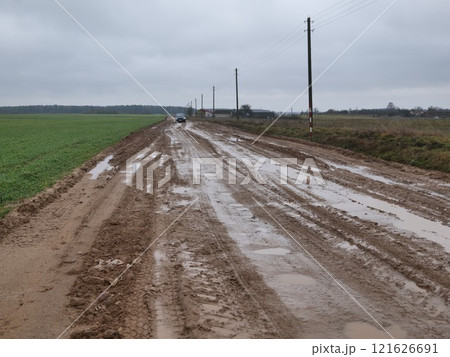 A muddy rural road, dotted with puddles, is lined with power lines and features an overcast gray sky overhead A muddy rural road, dotted with puddles, is lined with power lines and features an overcast gray sky overhead 121626691