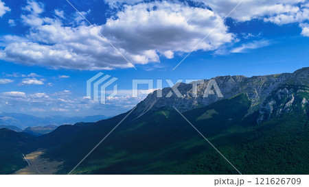 Majestic mountain range casting shadows over green valley under blue sky Majestic mountain range casting shadows over green valley under blue sky 121626709