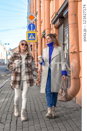 two young girls are walking along the city street, drinking coffee, talking two young girls are walking along the city street, drinking coffee, talking 121627174
