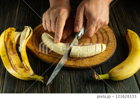Hands skillfully cut a perfectly ripe banana on a rustic wooden board. Fresh bananas surround the cutting area, hinting at a delicious treat to come 121628133