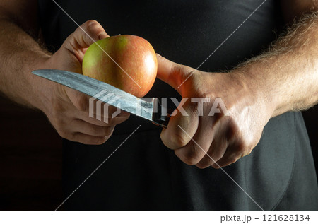 A pair of strong hands holds a shiny red apple while expertly gripping a gleaming knife. The warm, dim light creates an inviting atmosphere, enhancing the concentration on food preparation 121628134