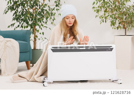 Young woman in socks with warm plaid near electric heater at home. Concept of heating season. Young woman in socks with warm plaid near electric heater at home. Concept of heating season. 121628716