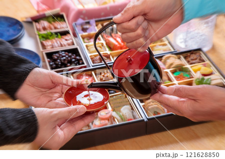 お節料理を背景に新年の祝い酒を注ぐ親子シーン お節料理を背景に新年の祝い酒を注ぐ親子シーン 121628850