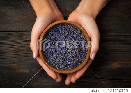 Hands gently holding a wooden bowl filled with dried lavender flowers on a rustic wooden surface 121630396