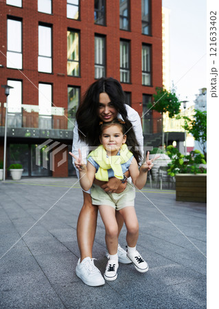 A woman playfully embraces a child, making peace signs in a modern urban setting during daylight 121633402