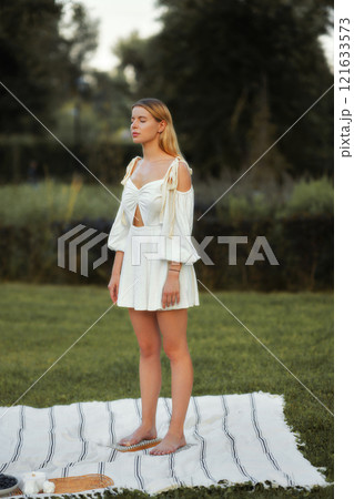 Model poses gracefully in a white dress on a blanket in a grassy area 121633573