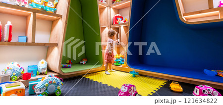 Child playing in a colorful playroom with shelves full of toys and cushioned seating areas 121634822