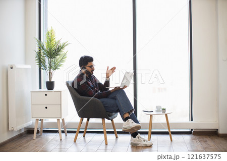 Indian man wearing headphones works remotely on laptop in home setting. Casual attire suggests comfort as he communicates. Natural light and plants add to relaxed ambiance. 121637575