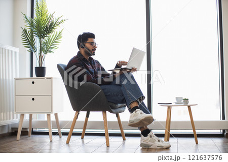 Indian male in late 20s using laptop in home office. Engaged in video call, wearing headset, gesturing okay symbol. Modern workspace with plants creates a cozy and productive atmosphere. 121637576