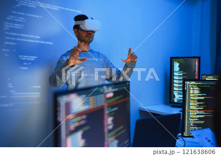 developer uses a VR headset to interact with projected interface designs, while colleagues discuss and code on screens, highlighting the integration of virtual reality and UI/UX design in modern developer uses a VR headset to interact with projected interface designs, while colleagues discuss and code on screens, highlighting the integration of virtual reality and UI/UX design in modern 121638606