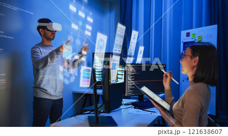 developer uses a VR headset to interact with projected interface designs, while colleagues discuss and code on screens at office 121638607