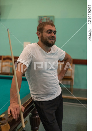 A handsome man stands in the billiard room holding a cue in his hands, leaning on the table and waiting for his turn. A group of friends decided to relax and play billiards on a weekend evening. A handsome man stands in the billiard room holding a cue in his hands, leaning on the table and waiting for his turn. A group of friends decided to relax and play billiards on a weekend evening. 121639263