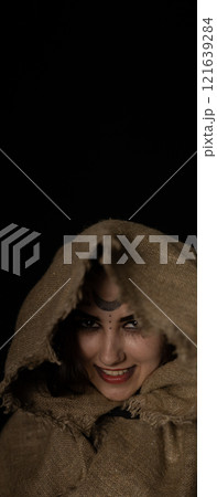 vertically. A grinning witch peeks out from under the hood on Halloween night. A beautiful witch with a hood thrown over her head smirks while looking at a vamer on a black background. Close-up. 121639284