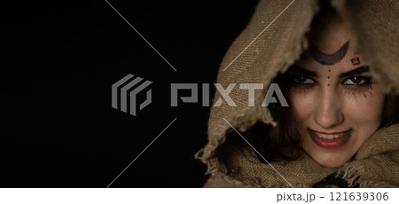 A grinning witch peeks out from under a hood on Halloween night. Copy space. Banner, flyer. A beautiful witch with a hood thrown over her head smirks while looking at a vammer on a black background. 121639306