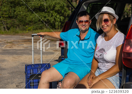 A man and a woman with a suitcase are preparing to go on a trip by car. They look at the camera. A beautiful couple of travelers in sunglasses sit on the open trunk of a car and look at the camera. A man and a woman with a suitcase are preparing to go on a trip by car. They look at the camera. A beautiful couple of travelers in sunglasses sit on the open trunk of a car and look at the camera. 121639320
