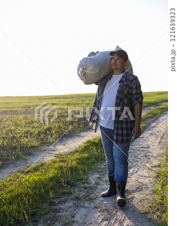 In work clothes, a man stands in a field with a bag on his shoulder at sunset. A worker in the field harvests in autumn. A man carries a bag over his shoulder into the car. In work clothes, a man stands in a field with a bag on his shoulder at sunset. A worker in the field harvests in autumn. A man carries a bag over his shoulder into the car. 121639323