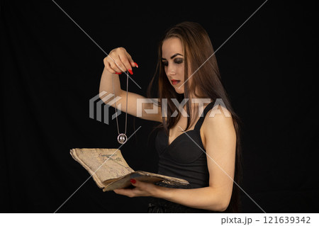 On Halloween, the witch performs a ritual with a book and a talisman. A beautiful female witch reads fortunes from an ancient book, holding a talisman in her hand. Halloween divination concept. 121639342