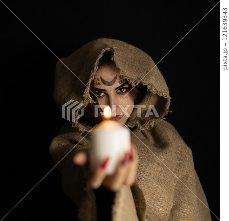 On Halloween night, a hooded witch holds out a burning candle from the darkness. The witch conjures in the evening on halloween over the flame of a candle. Halloween holiday concept. 121639343