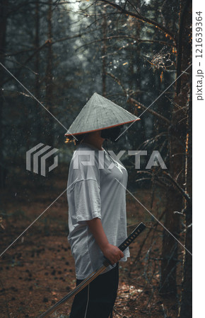 A woman wearing a straw hat and mask, depicting a samurai, stands in a forest, holding a katana at her side. The image evokes a sense of mystery and adventure. Woman with a katana. 121639364