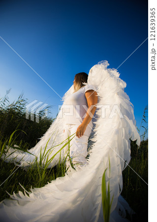 A woman with angel wings stands in a field, looking up at the sky. A magical scene that evokes hope and freedom. The sunlight bathes the angel in a bright glow, highlighting her purity and innocence. 121639365