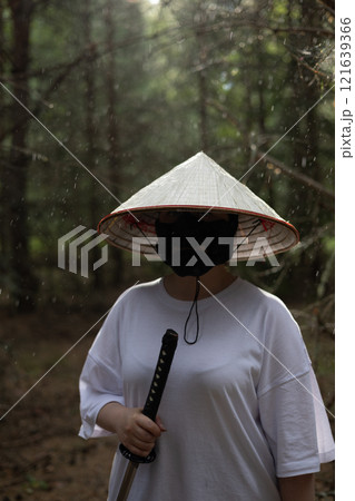 A man in a white shirt and a conical Asian hat holds a katana sword while standing in a forest. Rain falls on the trees in the background. A warrior with a katana stands in the forest in the rain. A man in a white shirt and a conical Asian hat holds a katana sword while standing in a forest. Rain falls on the trees in the background. A warrior with a katana stands in the forest in the rain. 121639366